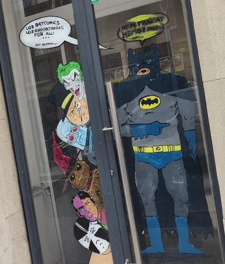 Una pintura en la puerta de cristal de una tienda muestra a Batman señalando hacia un grupo de villanos apilados, incluyendo al Joker y al Pingüino. Hay globos de texto humorísticos en español sobre la ubicación de los cómics y el cierre del local.
