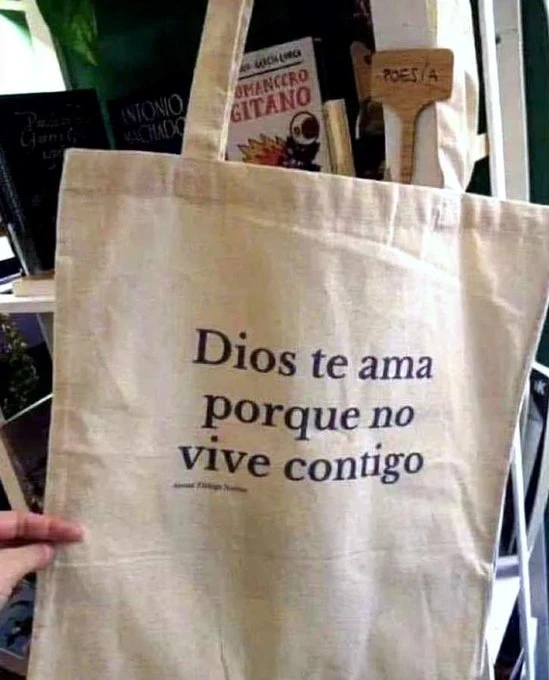 Bolsa de tela de color crema con la frase impresa "Dios te ama porque no vive contigo". Al fondo se aprecian varios libros de poesía en una estantería.