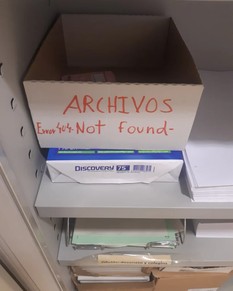 Una caja de cartón blanca etiquetada como 'ARCHIVOS' con la broma 'Error 404 - Not Found-' escrita debajo en rojo. La caja se encuentra en una estantería de oficina sobre un paquete de papel para imprimir.