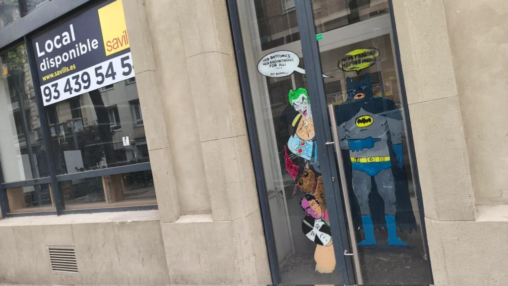Fachada de un local comercial disponible para alquilar con ilustraciones de Batman y sus villanos pintadas en el cristal de la puerta. Se observa un cartel de Savills indicando la disponibilidad del local.