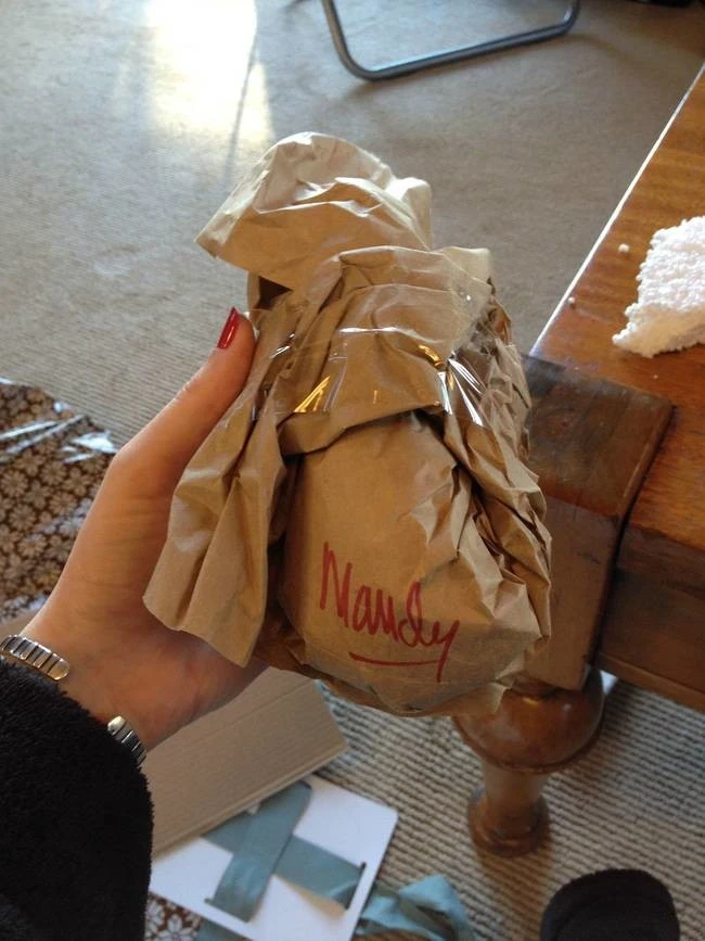 Una mano con uñas rojas sostiene un paquete envuelto en papel de estraza arrugado con el nombre 'Nandy' escrito en rojo. El paquete está sellado con cinta adhesiva transparente.