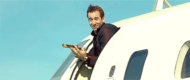 El actor Nicolas Cage asoma por la puerta de un avión, con una amplia sonrisa maníaca y sosteniendo una pistola dorada. La imagen animada muestra cómo su sonrisa se ensancha sutilmente y la pistola se mueve ligeramente hacia arriba.